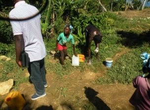 A young girl with braided hair carries a jug of water on her head in a rural area of Haiti.